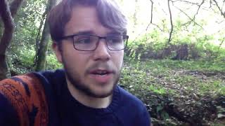 An 18 year old boy/man sits in the woods looking anxious, looking to camera.