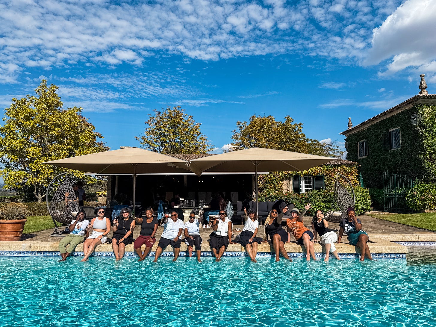 a sparkling blue pool with a large cabana house behind it and a row of women sitting smiling drinking wine with their feet in the pool.