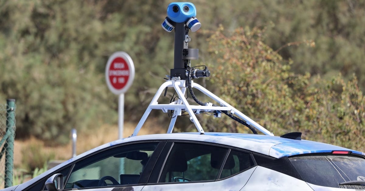 A Google Street View car with a large blue multi-lens camera mounted on top drives past a road sign and trees.