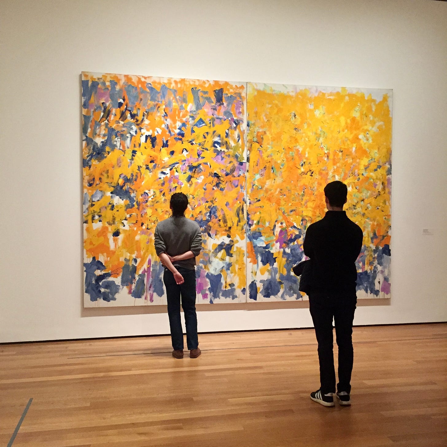 Two museum-goers gaze up at a large canvas: an abstract painting composed of yellow and blue marks