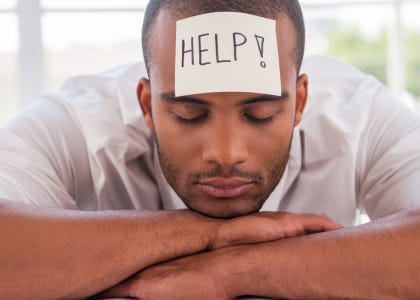 Man leaning on a table with his arms crossed resting his head on his hands with his eyes closed. He has a sticky note on his forehead that says "help!: