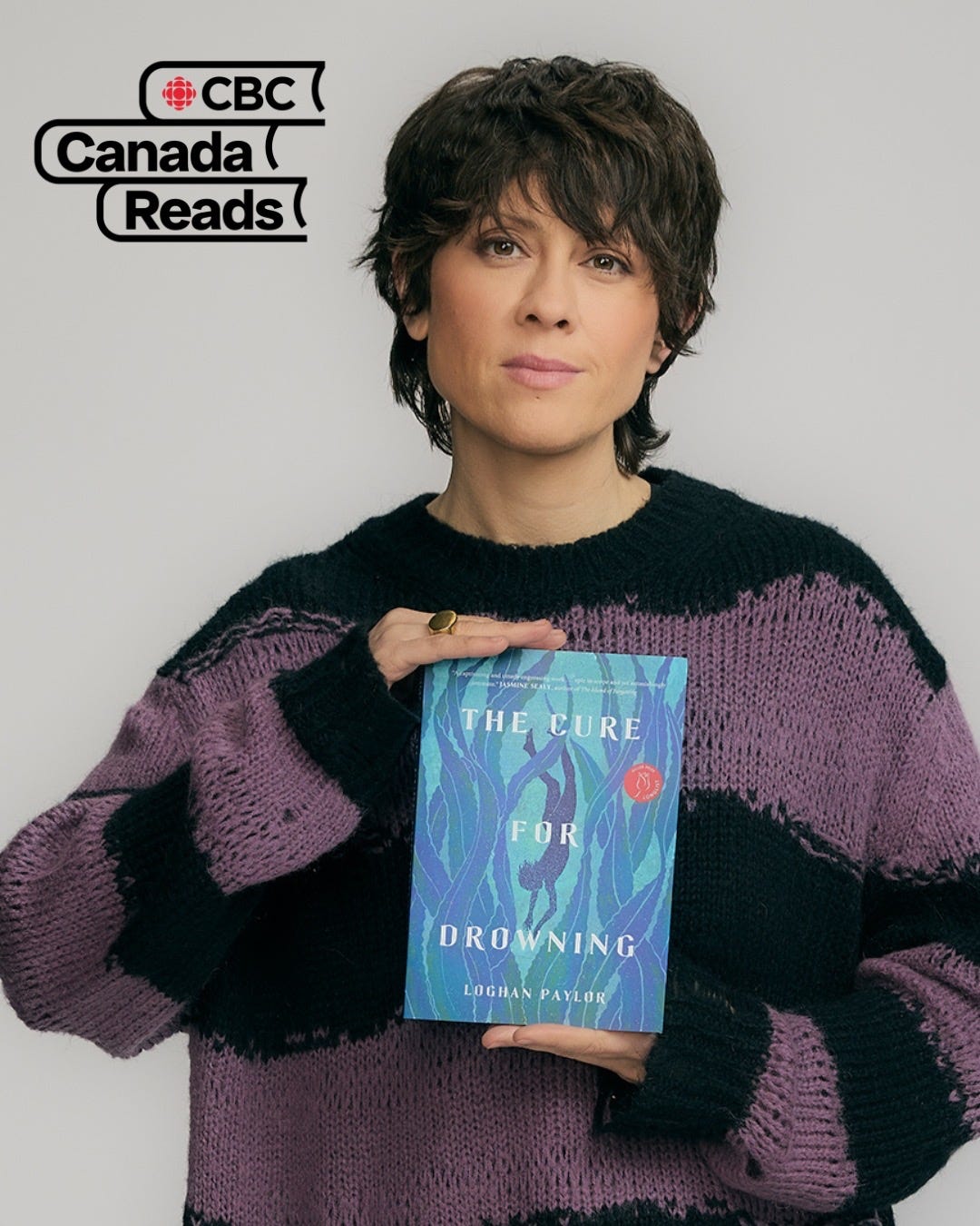 A picture of Tegan Quin wearing a black and purple shirt holding the book "The Cure for Drowning" by Loghan Paylor. A picture of Tegan Quin wearing a black and purple shirt holding the book "The Cure for Drowning" by Loghan Paylor.
