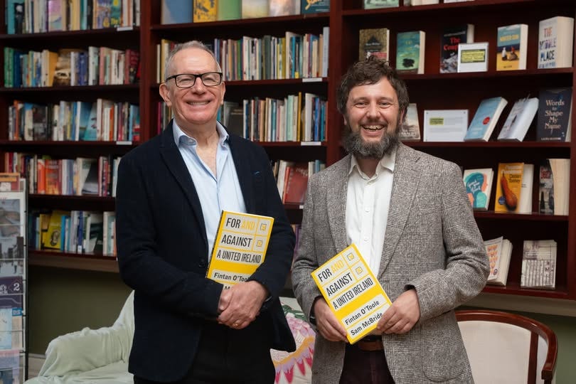 Two men stand in front of bookshelves, each holding a yellow book, smiling at the camera.