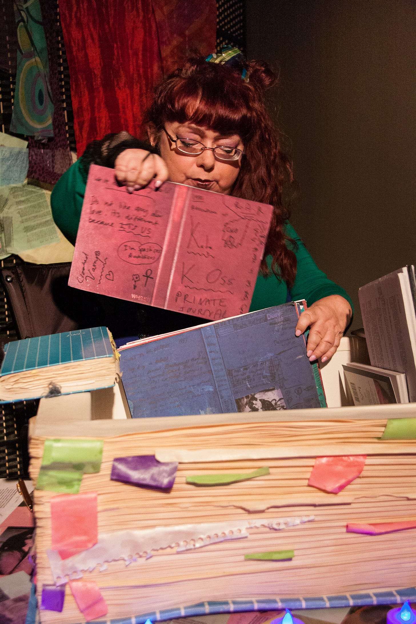 An image of Penny in dark, rich tones, dark red velvet draped behind her, with a spotlight effect highlighting her face. Penny has dark red hair in a rough style, sits in her wheelchair and wears a dark green cardigan. She’s on stage performing in Lost in Spaces. The front of the image highlights a prop in the shape of a huge replica of one of her diaries. The original diary, slightly bigger than a paperback book, is to the left, blue with white stripes, bulging pages with tattered paper hanging out. The replica is as big as a dining table, and recreates the design of the original, the main cover acting as a lid which opens like a giant book to reveal a space with the diaries inside. These have different coloured covers, which Penny is holding. They are scribbled on with crazy, punky, handwritten notes and titles.