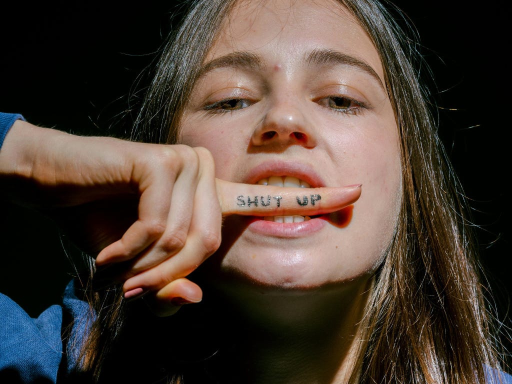Close-up van een vrouw met een vinger voor haar mond waarop ‘SHUT UP’ staat, met daarover in witte letters het woord Gatekeepers.