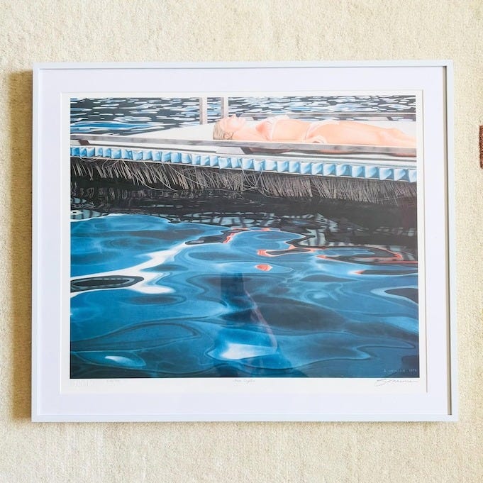 May include: A framed art print featuring a person floating in a pool. The water is a vibrant blue with light reflections. The frame is white, and the artwork is displayed against a cream-colored rug with colorful square accents.