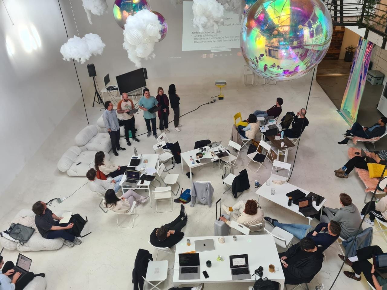 A photo taken from above of people in a large white room - they are sitting on chairs and around desks and talking informally. 