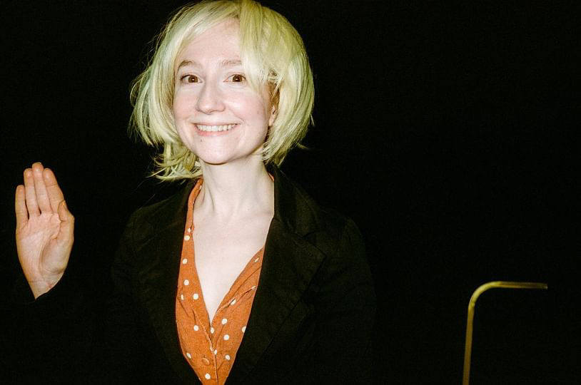 Female comedian in short blonde wig smiles at camera with awkward hand wave Female comedian in short blonde wig smiles at camera with awkward hand wave