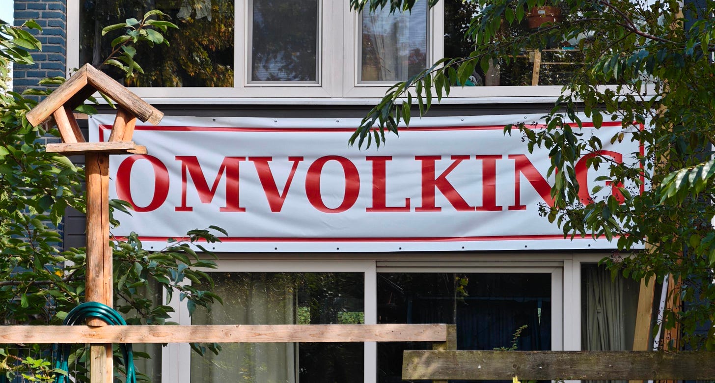 Gevel van een rijtjeshuis met tussen de ramen een wit spandoek met in grote rode letters OMVOLKING. Ervoor een houten vogelhuisje, een opgerolde tuinslang en een schutting. Gevel van een rijtjeshuis met tussen de ramen een wit spandoek met in grote rode letters OMVOLKING. Ervoor een houten vogelhuisje, een opgerolde tuinslang en een schutting.