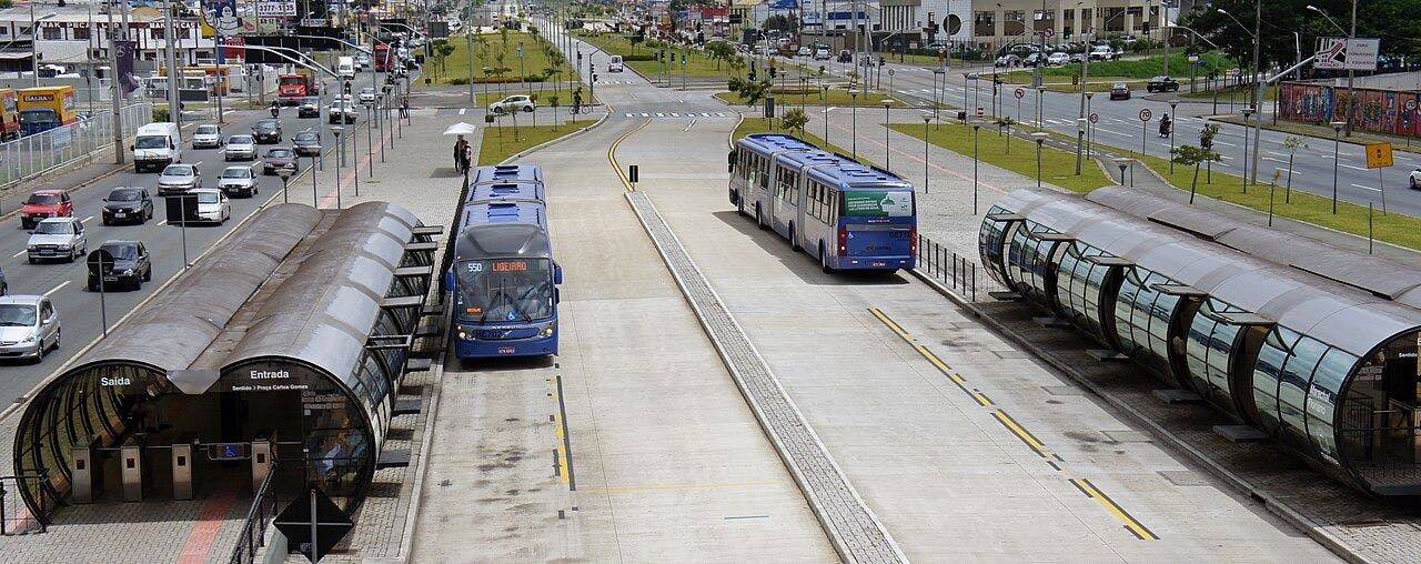 two "stations" shaped like double-tubes, either side of a road at which a pair of articulated buses are about to arrive or leave two "stations" shaped like double-tubes, either side of a road at which a pair of articulated buses are about to arrive or leave