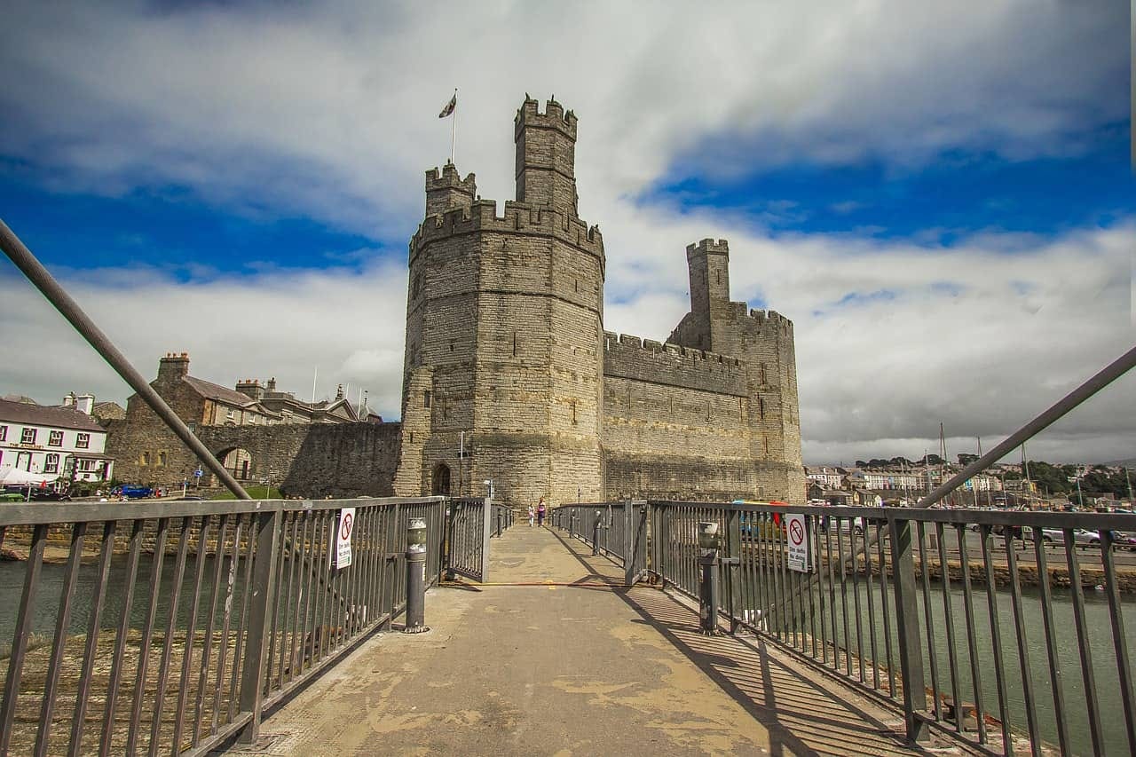 A ground-level view looking across a pedestrian footbridge to the massive stone walls and polygonal towers of Caernarfon Castle, with the River Seiont visible below