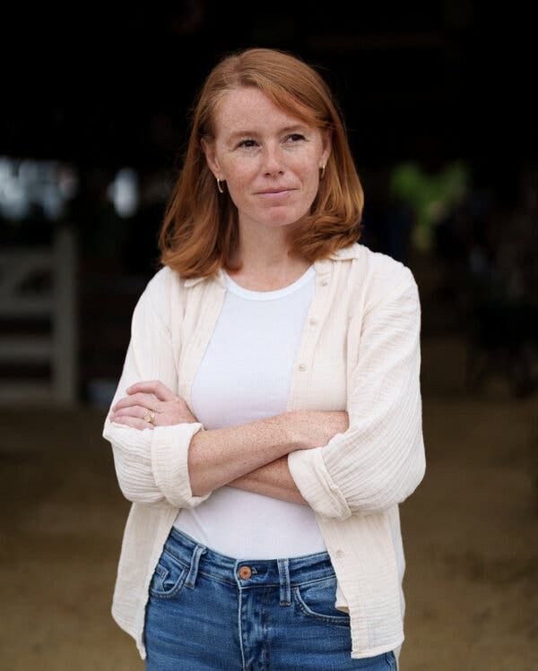 A woman with red shoulder-length hair and gold hoop earrings posing for a portrait outside with her arms crossed. 