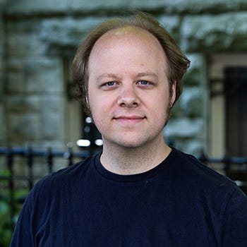 Tim's headshot. He's quite dashing in a blue t-shirt that brings out his eyes, the wind feathering his youthful hair, standing in front of a gothic church.