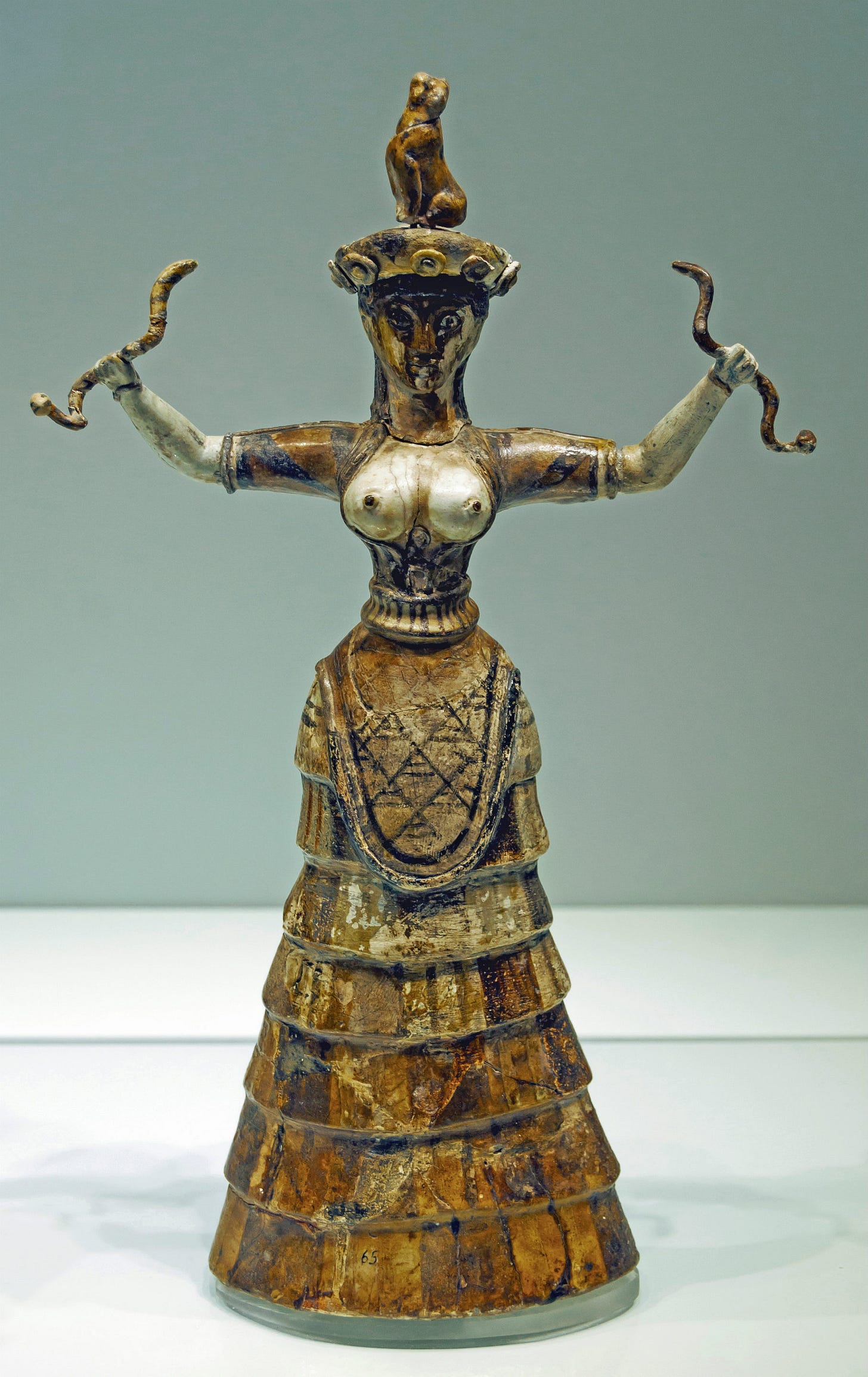 The Minoan Snake Goddess faience figurine, made around 1600 BCE in Knossos, as displayed in the Heraklion Archaeological Museum. She is wearing a long flounced skirt, an apron, an open-front short-sleeved top that shows her breasts, and a beret type hat. A cat sits on top of her hat. She is holding up a writhing snake in each hand.