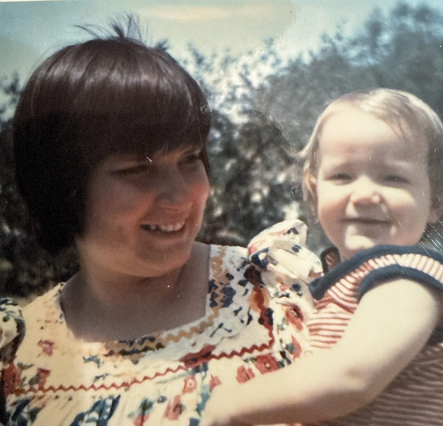 A young mother with dark brown hair in a bob with bangs smiles at the baby she is holding.