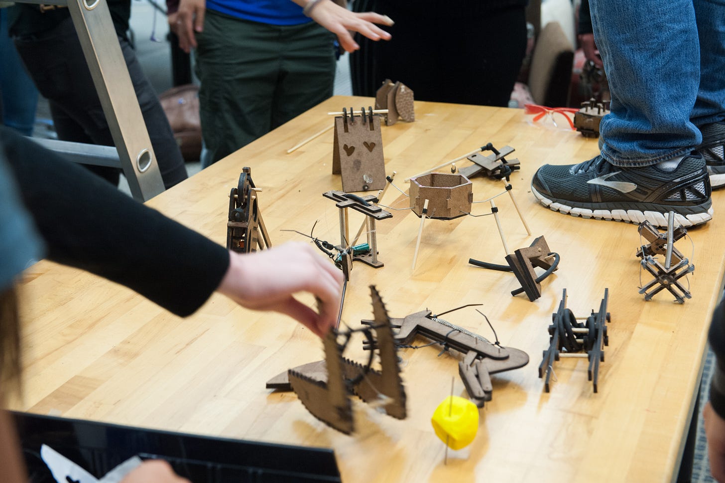 A table top holds a dozen small gadgets built of particle board and spring kits, all built to maximize a hopping mechanism. Students hands reach into the frame, and one student stands on top of the table, only feet and legs visible. A table top holds a dozen small gadgets built of particle board and spring kits, all built to maximize a hopping mechanism. Students hands reach into the frame, and one student stands on top of the table, only feet and legs visible.