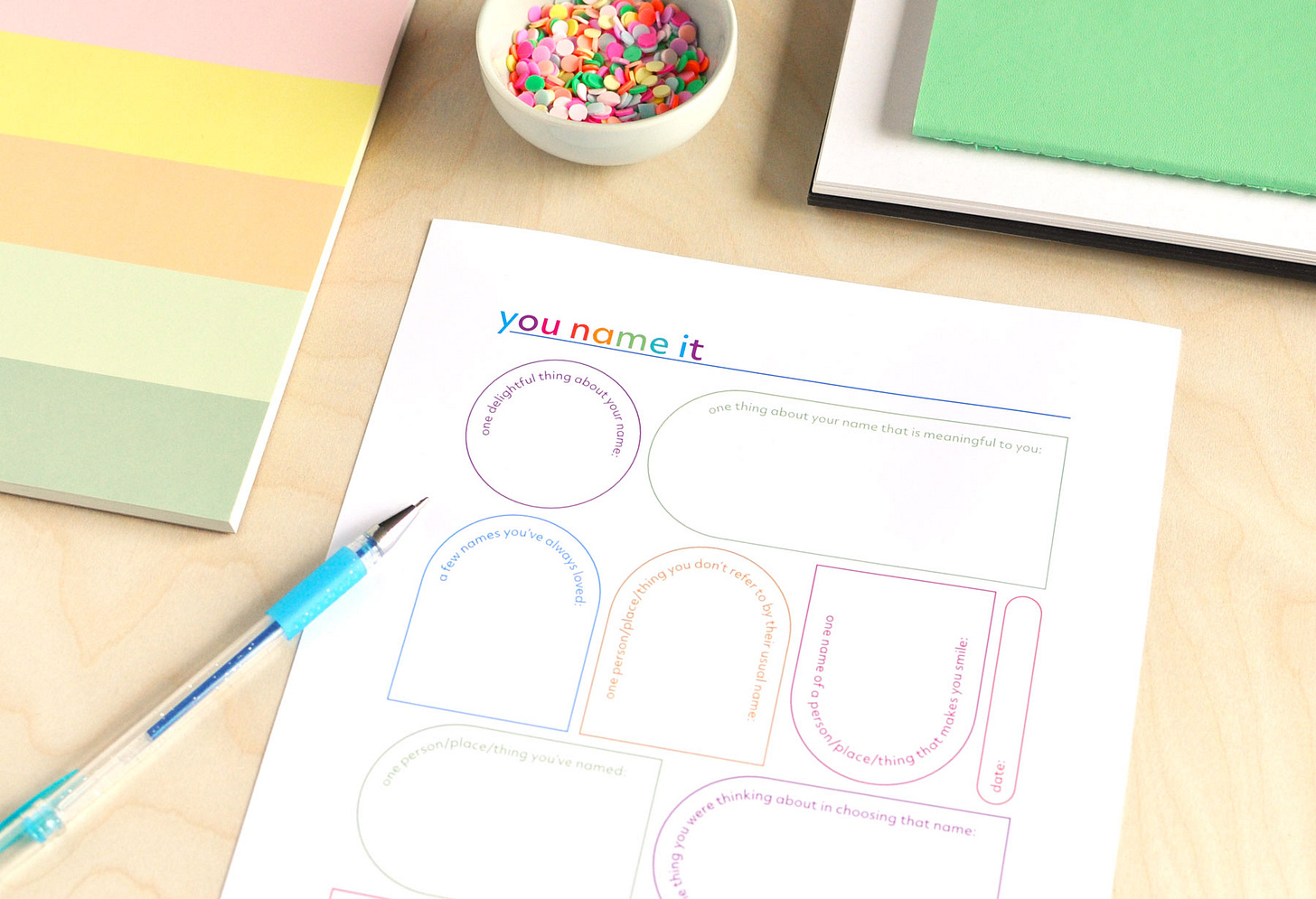 A colourful journal page titled You Name It, filled with journal prompts about the names we give things. Shown on a wood background with blue pen and colourful notebooks.