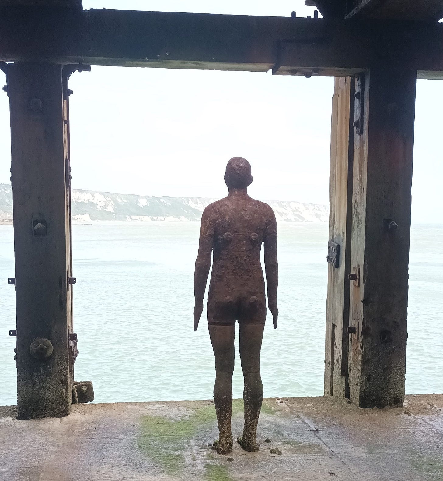 Another Time XVIII, Anthony Gormley, a weathered bronze life size male form, looking out to the sea and cliffs beyond