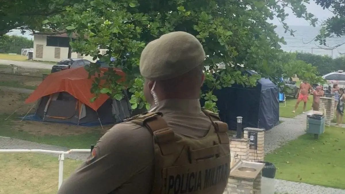 View from behind a uniformed military police officer wearing tactical gear, standing near a grassy campground with tents and temporary structures. Several people are visible in the background near the beach access area.
