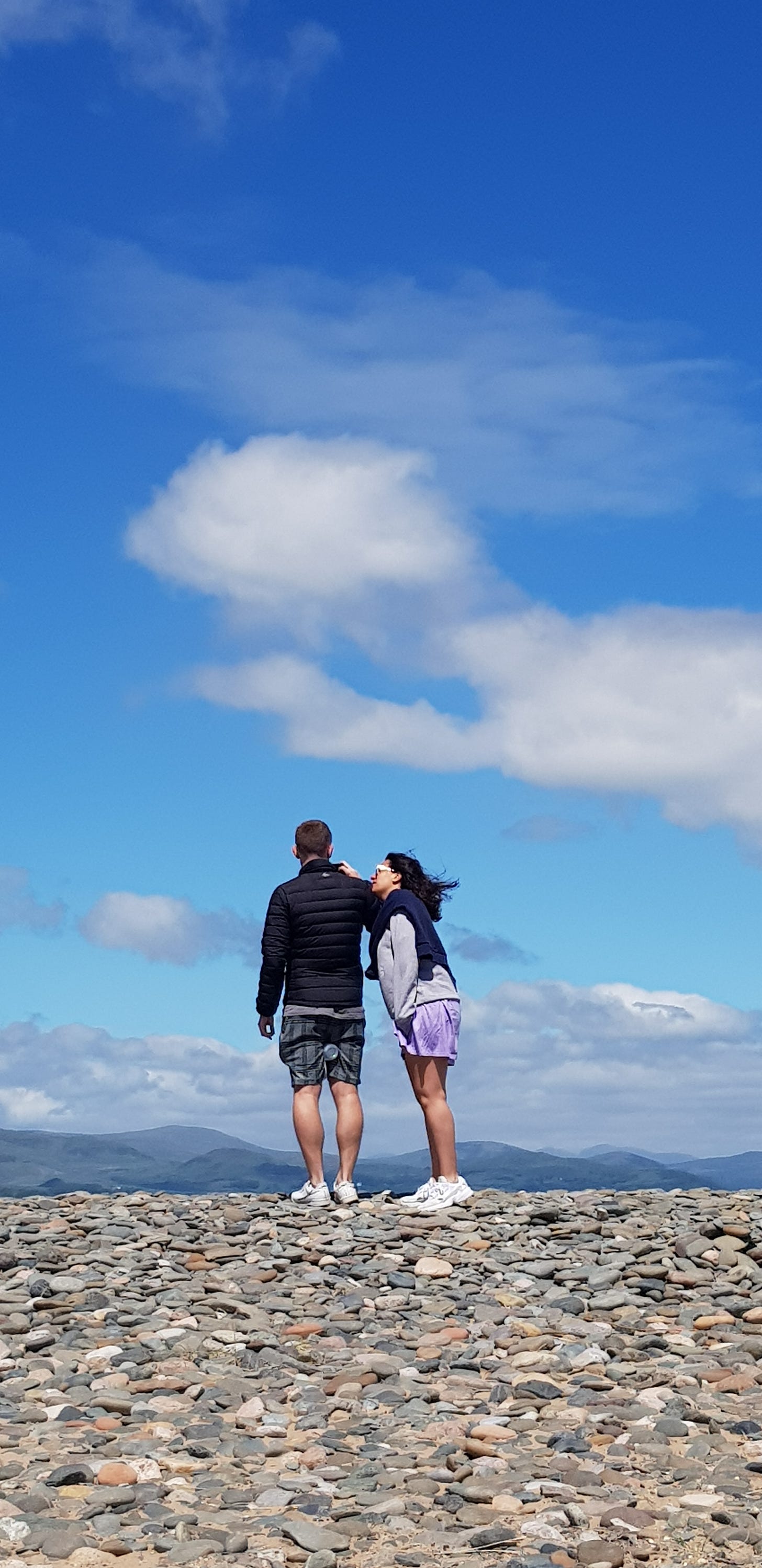 Taken from a distance a couple take in beautiful blue skies and cumbrian views. They're on a pebbly beach