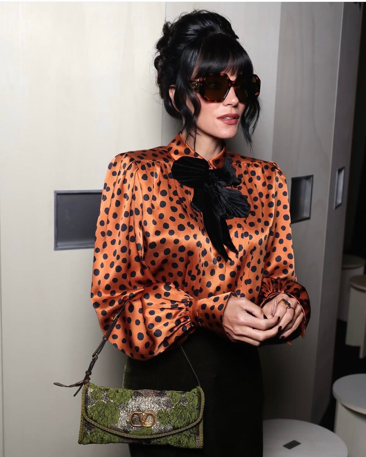 Lilly Allen standing with 60's bouffant hair do, dark glasses, wearing Valentino orange silk blouse with black polka dots and big pussy cat black bow tied at neck , she is carrying a green snake leather type handbag. It is at Paris Haute Couture Week 2026.