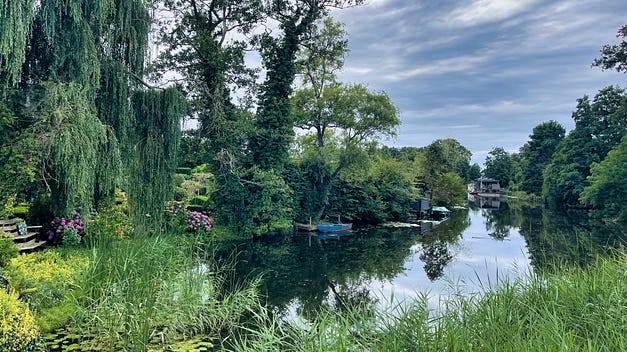 A quiet river, it looks more like a pond on this photo, with green trees and flowers on all sides. The blue sky is reflected on the water.
