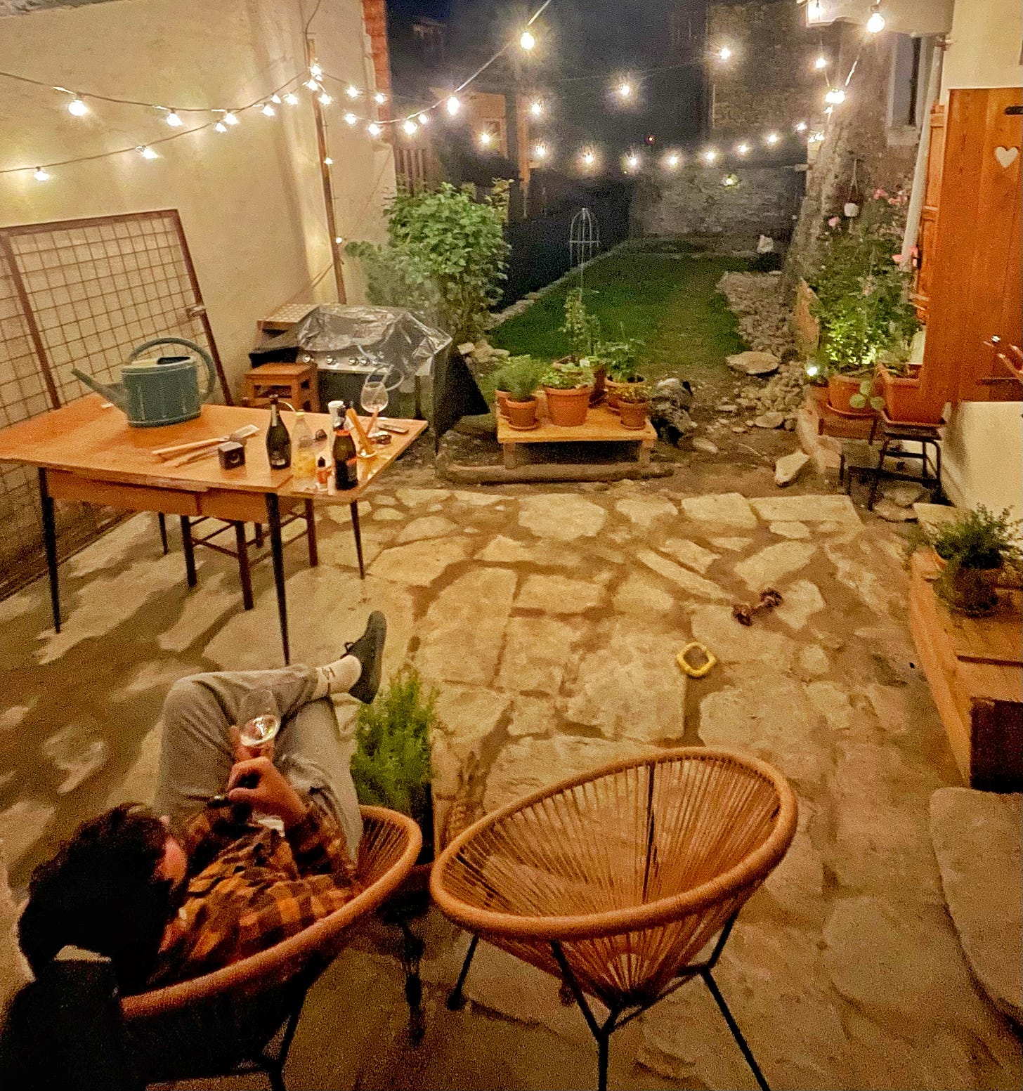 A romantic garden terrace, in the evening, with fairy lights, herbs growing in pots and a man sat relaxing in a chair