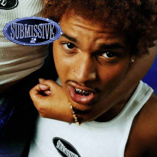 A close-up album-cover image shows a person looking up toward the camera with a direct, expressive gaze. They wear a white tank top, gold grillz, a nose piercing, and a gold bracelet. An arm from another person rests across their shoulder. A metallic oval logo reading ‘Submissive 2’ appears in the upper left corner, giving the image a bold, provocative aesthetic.