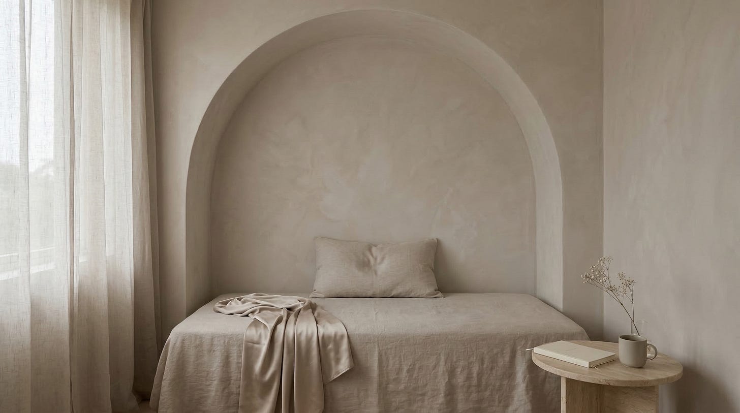 A linen daybed beneath a plaster arch alcove with a single pillow and a rumpled matte champagne satin throw, beside a small round side table holding a closed cream journal, a ceramic mug, and dried baby's breath in a glass vase, with soft light filtering through sheer linen curtains