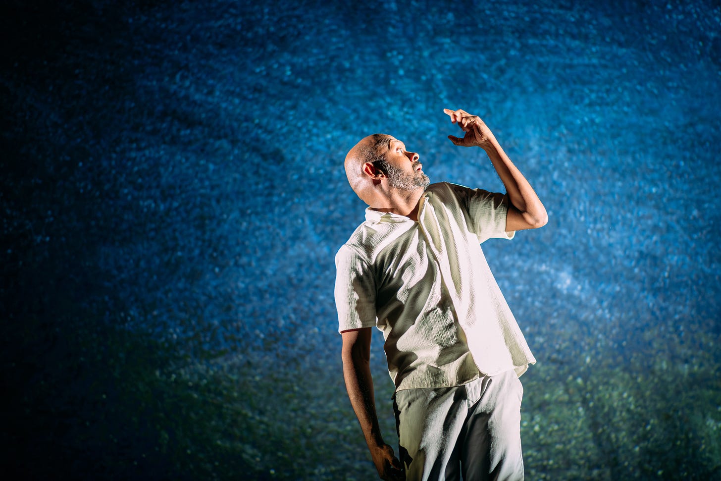 Performer Ramesh Meyyappan in a white shirt and grey trousers. Ramesh is stood up facing the left towards his left hand which is held in front of his face with curved fingers. The back wall is full of galaxy - like projections. Photo taken by Miha ela Bodlovic