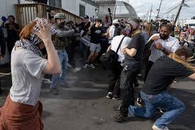Woman with brown hair wearing a black top with multicolored scarf; protesters and ICE agents; man with brown hair wearing a white dress shirt, ti, and black blazer; man with short black hair wearing a salmon-colored turtleneck and black blazer; man with gray hair wearing a white dress shirt, tie and black blazer.