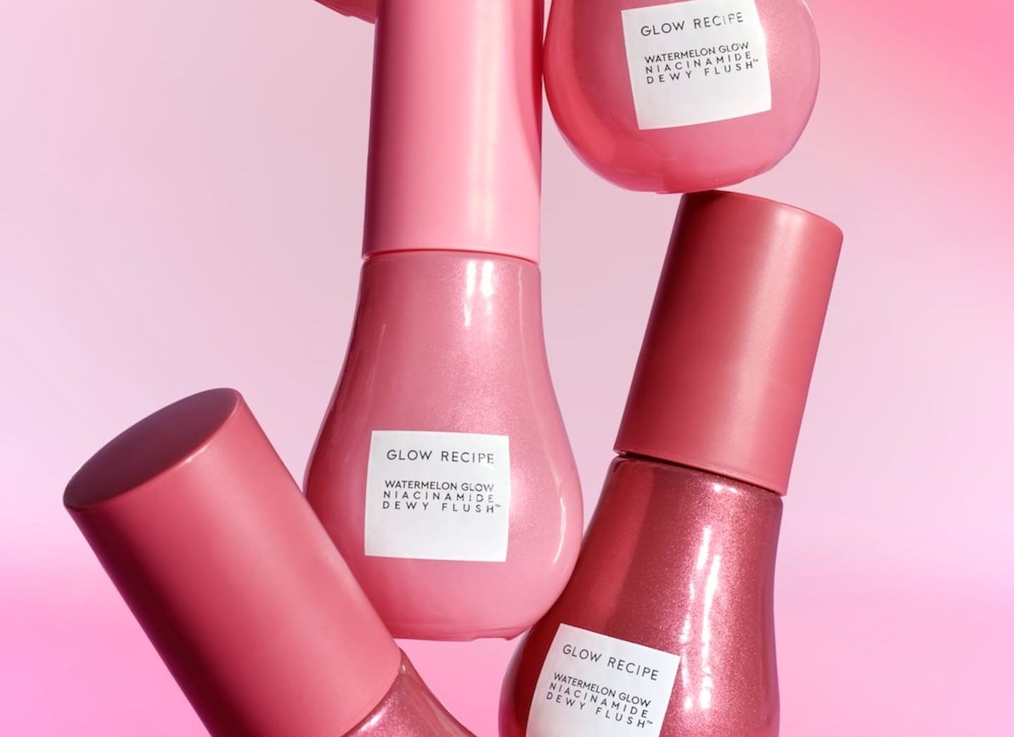 On a pink background, an artful cascade of blush bottles are displayed, their labels reading Glow Recipe Watermelon Niacinamide Dewy Flush.