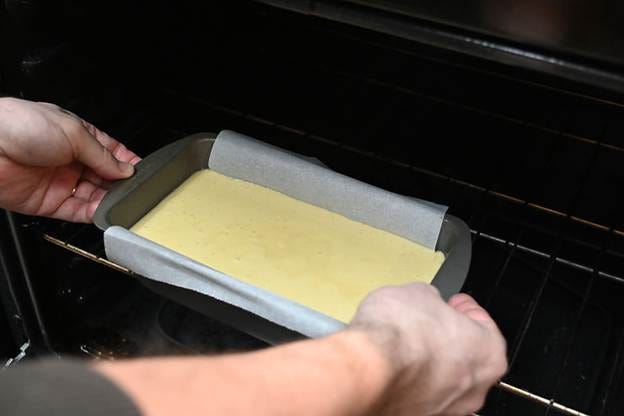 Hands place a parchment-lined baking tray with yellow batter into an oven, suggesting baking. Oven interior is dark. Hands place a parchment-lined baking tray with yellow batter into an oven, suggesting baking. Oven interior is dark.