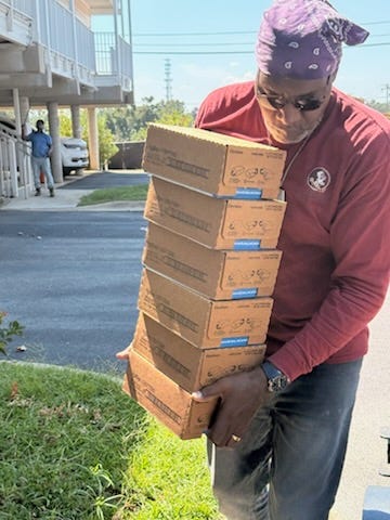 A person in a red shirt loads a red hand truck stacked with cardboard boxes, including several large Coffee mate French Vanilla cartons. Nearby, another person wearing a black T‑shirt with a yellow Batman logo and a camo cap stands beside a gray picnic table. The scene is an outdoor loading or side area next to a building with siding, columns, and concrete pavement.