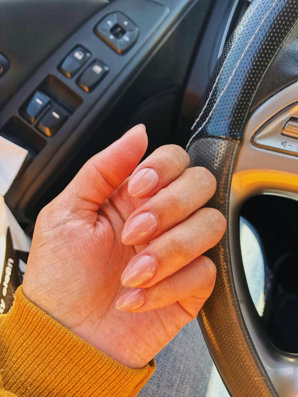 A hand showing off nude colored nails in a car, in front of a steering wheel.