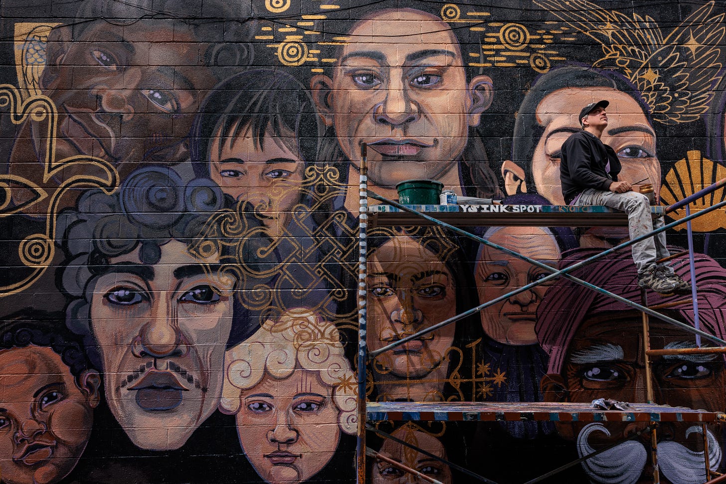 Pierre Nagley sits on scaffolding in front of an unfinished mural of overlapping portrait faces painted in black, gold, and muted tones on a brick wall.