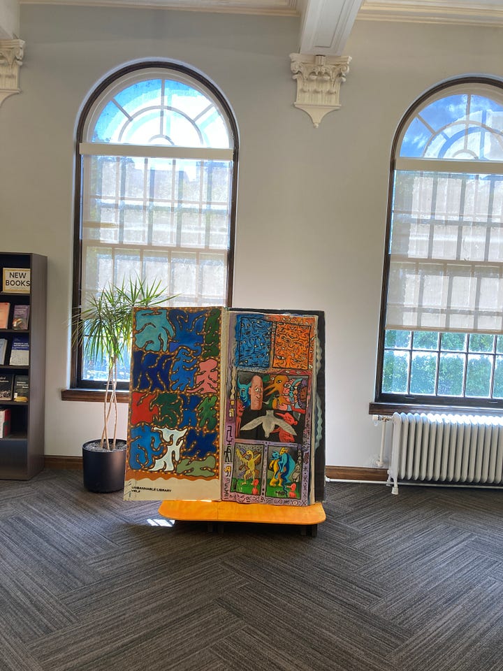 A big book with colorful pages sits on a display stand in front of a pair of windows. A painted, oversized book sits on a green display stand with library shelves visible behind it. A man stands next to an oversized book that is painted in red and green.