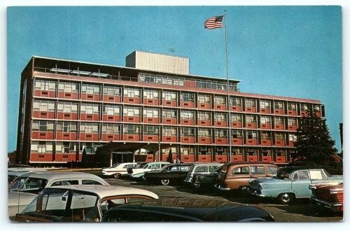 Postcard showing a community hospital from the 1960s, with a Plymouth Belvedere, Ford Galaxie, and Studebaker Lark Wagon in the parking lot, and an American flag fluttering in the breeze at the top of a very tall flagpole. The sky is cloudless and brilliantly blue.