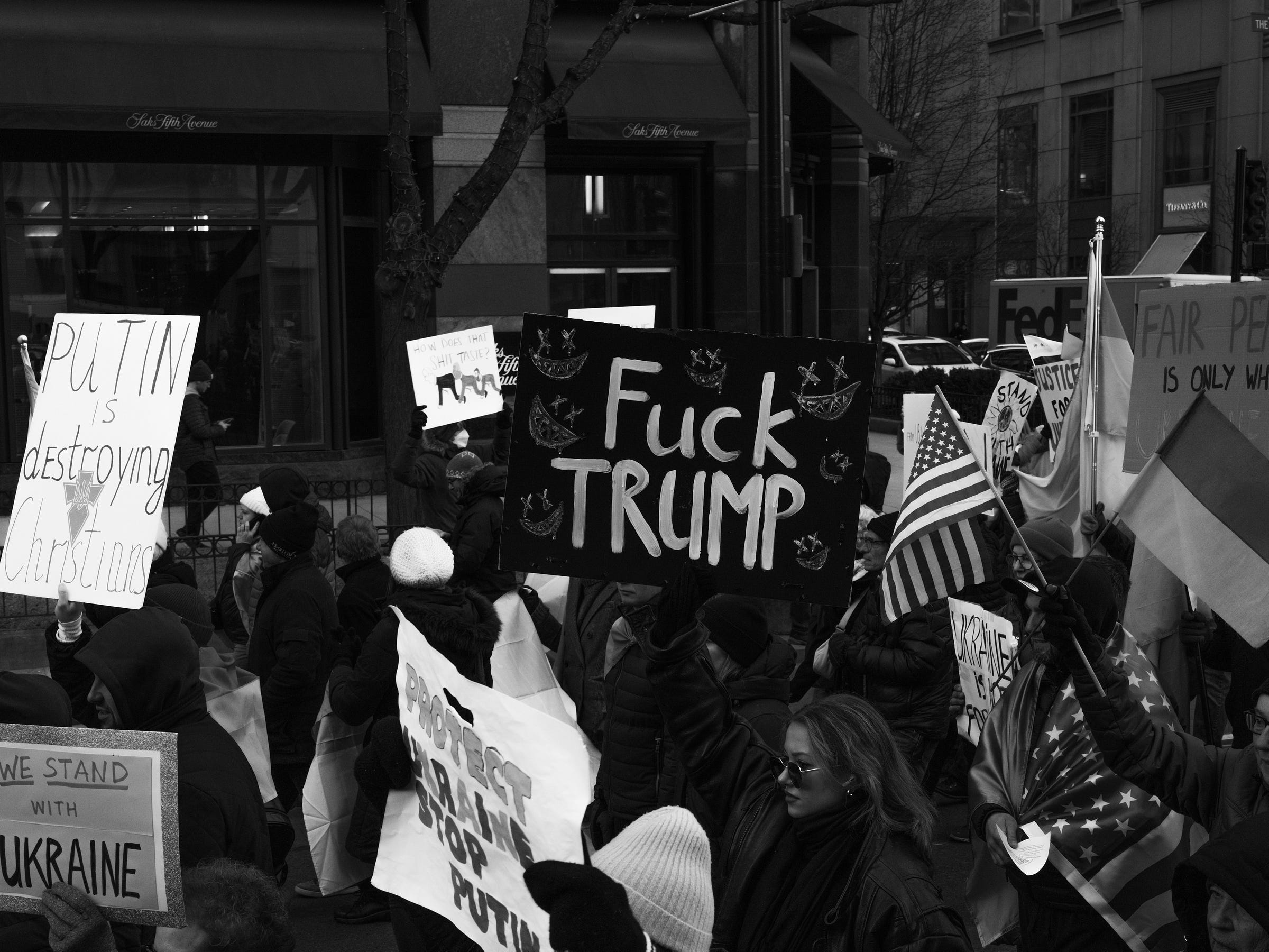A protest scene with demonstrators holding various signs and flags. One prominent sign reads "F*** TRUMP" with decorative elements, while another states "PUTIN is destroying Christians." American and Ukrainian flags wave among the crowd as people march in solidarity. The background features storefronts and city buildings. A protest scene with demonstrators holding various signs and flags. One prominent sign reads "F*** TRUMP" with decorative elements, while another states "PUTIN is destroying Christians." American and Ukrainian flags wave among the crowd as people march in solidarity. The background features storefronts and city buildings.