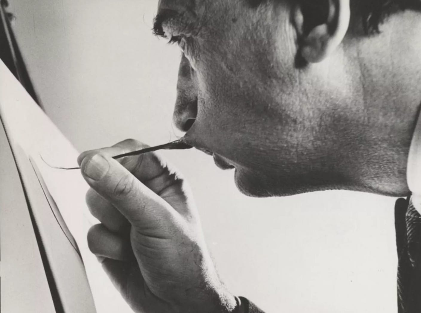 Black and white photograph of a man with a distinctive mustache using it to hold a thin paintbrush while drawing on a canvas. The close-up shot emphasizes the texture of his skin, the sharpness of his facial features, and the intensity of his concentration.