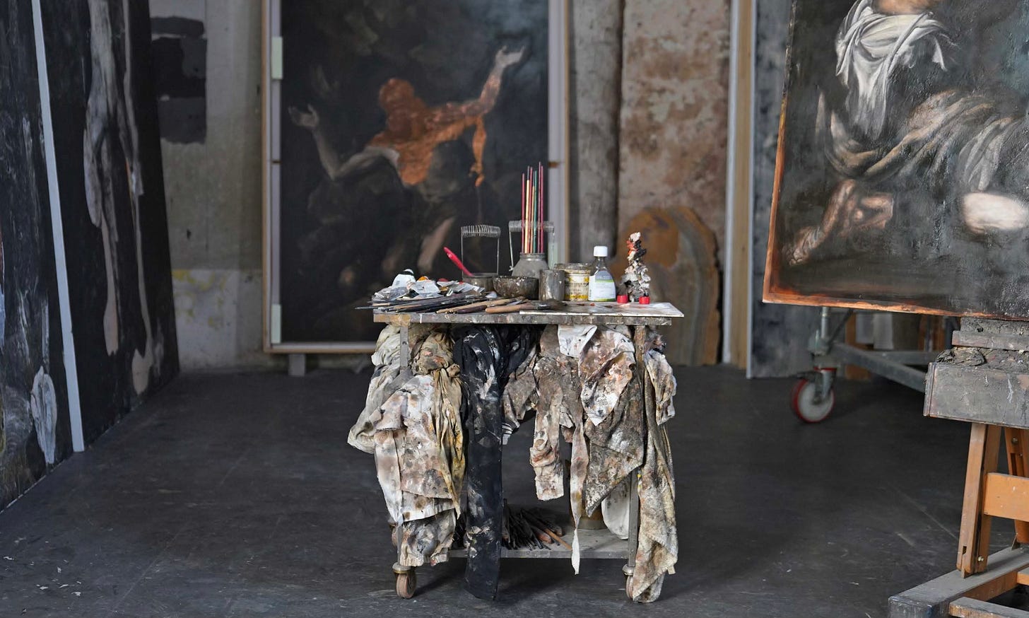 Studio of Italian painter Nicola Samorì: a paint-covered worktable at the centre of a large space, surrounded by large-scale dark figurative paintings on canvas. Photo by Mario Cresci, 2026. Curatorial project by Roberta Bertozzi.