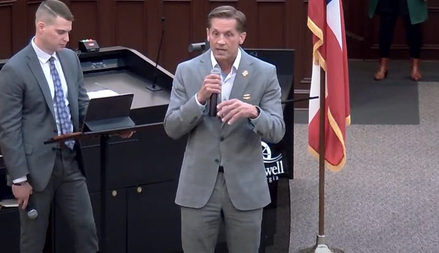 Video screenshot of Rep. Rich McCormick, wearing a light grey suit with no tie, standing in a public meeting room and speaking into a microphone. On the left side of the photo, an aide in a dark grey suit stands at a lectern with a tablet on it, holding a microphone at his side. Guess he's there to read audience members' questions?