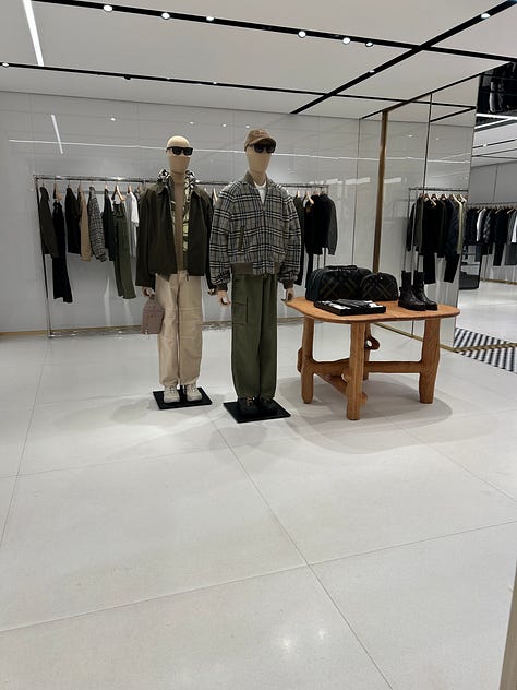 Three-photo gallery from an Intern Pierre primary channel-check store visit to a Burberry flagship, documenting the gap between the company's articulated heritage refocus narrative and the actual visual merchandising on the floor. Left photo: the women's handbag wall positioned at the storefront entry zone, with structured leather Burberry shoulder bags, hobo bags, and check-pattern canvas backpacks displayed on backlit white wall shelves above a glass jewelry display case and a beige suede storage bench. Center photo: a sparse men's ready-to-wear central display featuring two mannequins in muted olive and grey tailored outfits styled with bucket hats, flanked by half-empty wood-tone clothing rails and a minimalist wood bench, occupying the deep interior of the store far from the entrance. Right photo: men's outerwear and ready-to-wear racks in a back-of-store section, featuring a Burberry check-pattern bomber jacket with contrast sleeves, a red and beige check overshirt, and additional checked outerwear hanging on chrome rails with folded scarves and a leather portfolio bag arranged on a low display plinth. The sequence demonstrates the structural mismatch between the company's "hero categories take prominence" merchandising claim and the actual layout, where handbags and accessories anchor the entry zone, men's outerwear and trench coats require navigating deep into the store, and check-pattern saturation remains heavy across ready-to-wear and outerwear categories despite the public narrative of a quiet-luxury heritage repositioning. Channel-check evidence relevant to Burberry $BRBY.L investors evaluating the credibility of the current turnaround thesis and the alleged return to outerwear and scarves as the brand's anchor categories under CEO Joshua Schulman.