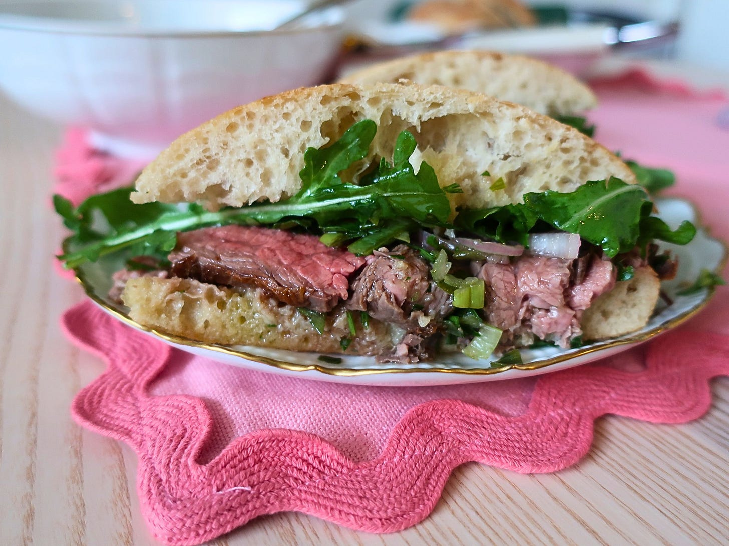 A halved steak sandwich with pink, medium-rare steak topped with bright green herbs and arugula on ciabatta rolls, all on a pink placemat.