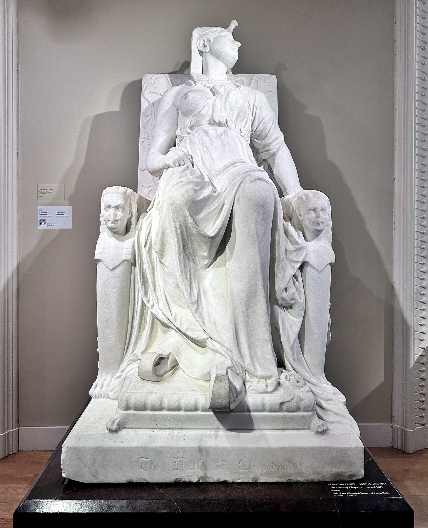 White marble statue of Cleopatra slumped on a throne, head tilted back and arms limp, with a carved head on each armrest. White marble statue of Cleopatra slumped on a throne, head tilted back and arms limp, with a carved head on each armrest.