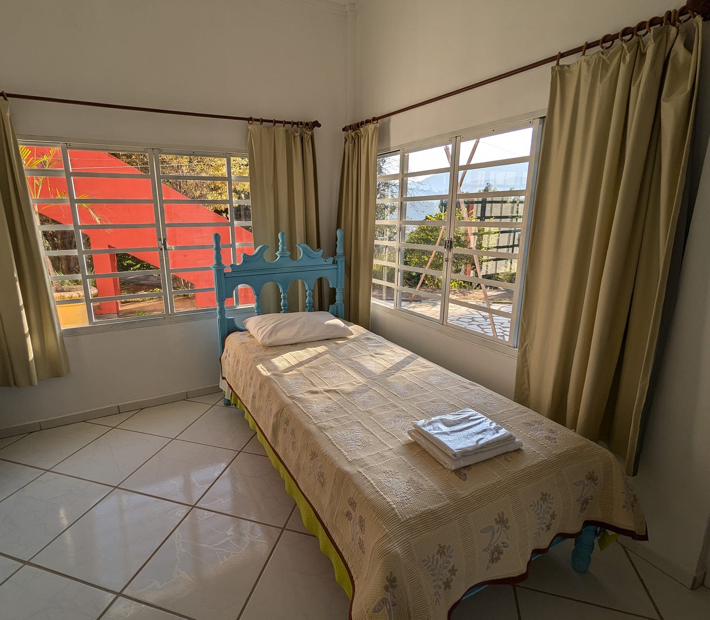 A simple bedroom in a Brazilian guesthouse with white tile floors and pale green curtains. A single bed with an ornate turquoise headboard sits between two large windows. Through the windows, red-tiled roofs, green hills, and wooden structures are visible. Folded white towels rest on the bed. Morning sunlight fills the room.