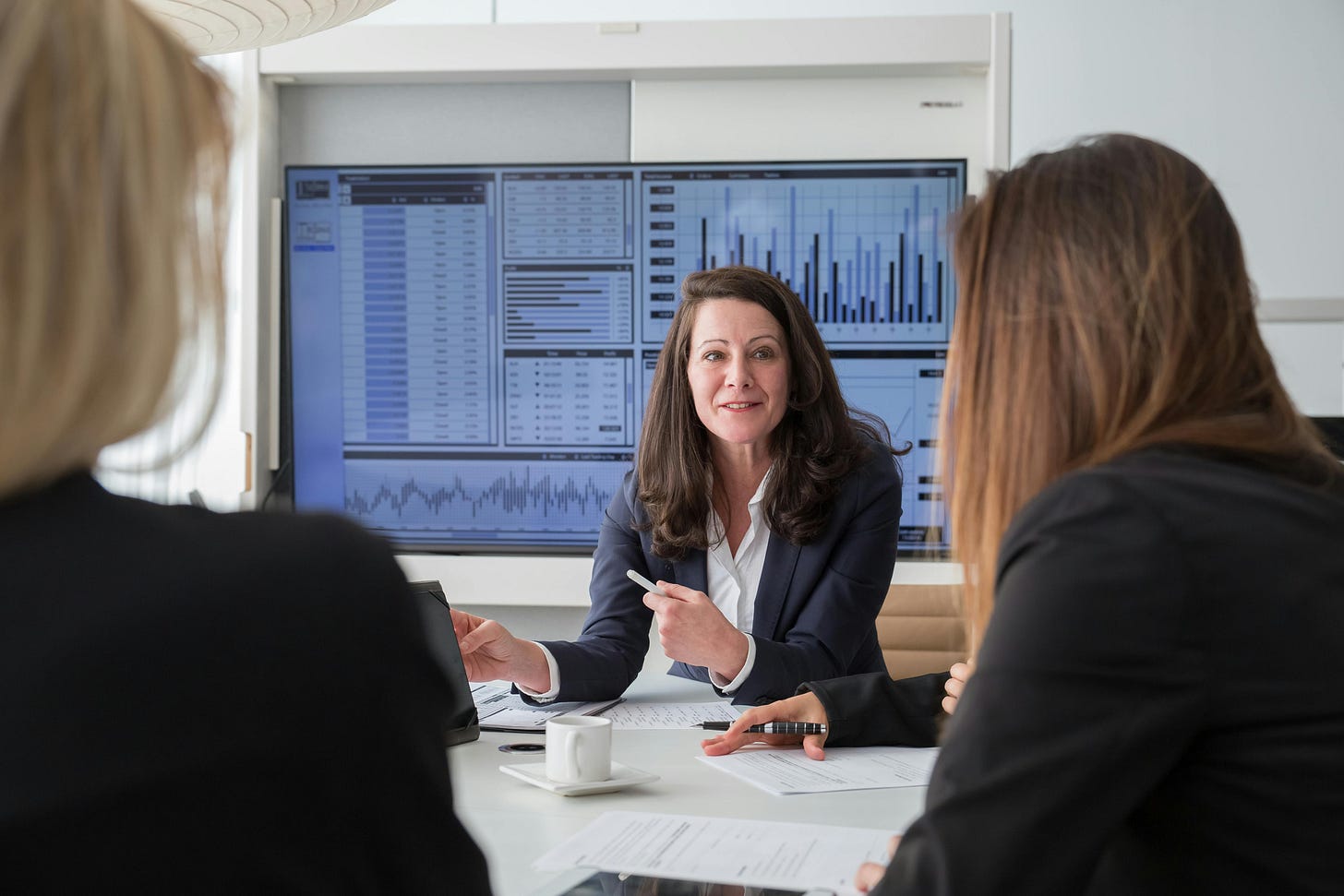 Businesswoman leading a boardroom meeting with investors, presenting financial charts on a large screen.