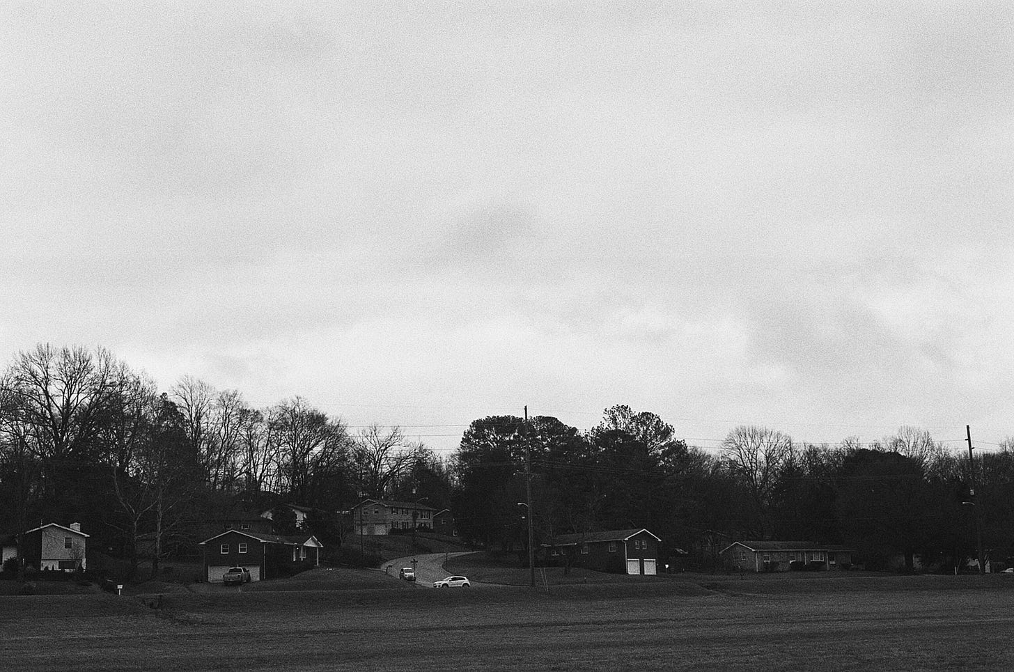 A black and white photograph of a row of houses across the road from a field. Tall trees tower behind them, but most of the photo is sky.