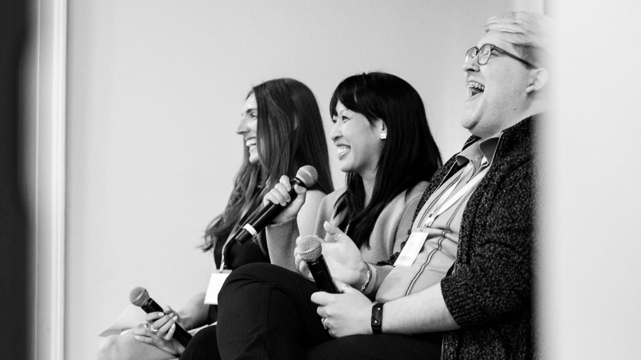 Black and white photo of three panelists seated and laughing while holding microphones. The two women and one man appear to be mid-discussion or reacting to something humorous during a live event or conference. All three wear name badges, suggesting they are speakers or moderators. The moment captures a lighthearted and engaging atmosphere.