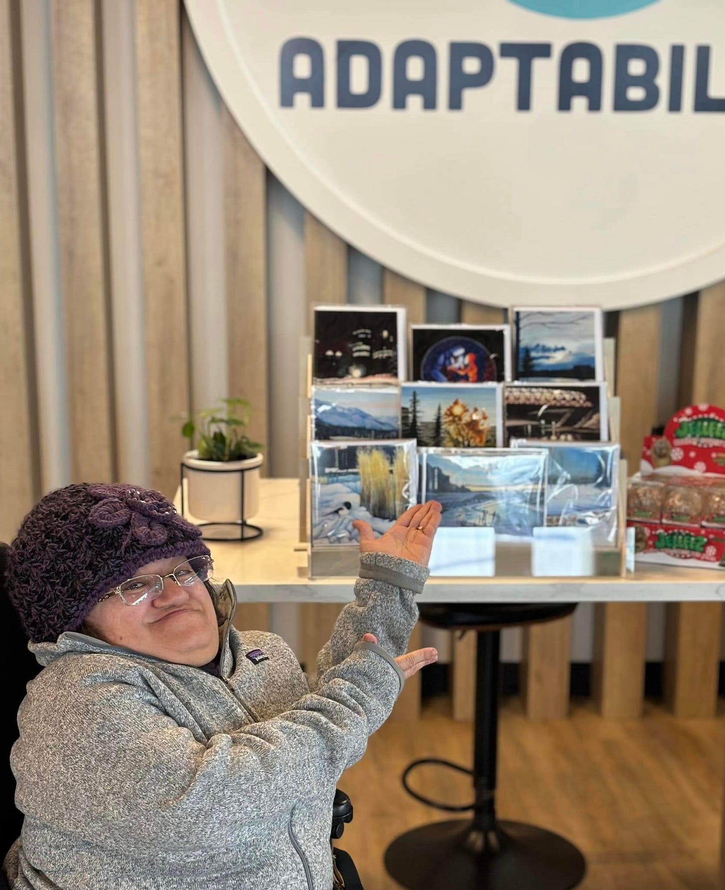 Athena wearing warm winter gear pointing to a display case with her greeting cards at the AdaptAbility store. Athena wearing warm winter gear pointing to a display case with her greeting cards at the AdaptAbility store.